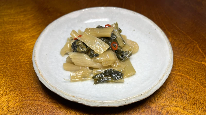 秩父の郷土食・しゃくし菜（杓子菜）の塩漬けをレシピ通りに調理し、唐辛子と出汁で味付けした油炒めを小鉢に盛りつけたもの。つやのある緑色の葉と赤い唐辛子が彩りよく、家庭で作る素朴な郷土の味わいが伝わる一枚